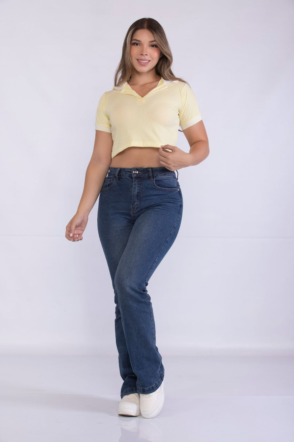 Mujer sonriente modelando el crop top polo Caelia en color amarillo pastel con cuello y ribetes blancos en contraste, combinado con vaqueros de cintura alta sobre un fondo blanco.